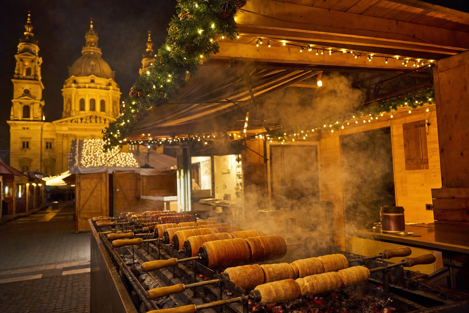 Budapest Christmas market stall at night, rows of golden chimney cakes rotating over glowing coals with steam rising into cold blue air, strings of warm fairy lights above wooden stall roofs, the lit dome of St. Stephen's Basilica soft in the background, festive winter mood, no people prominent