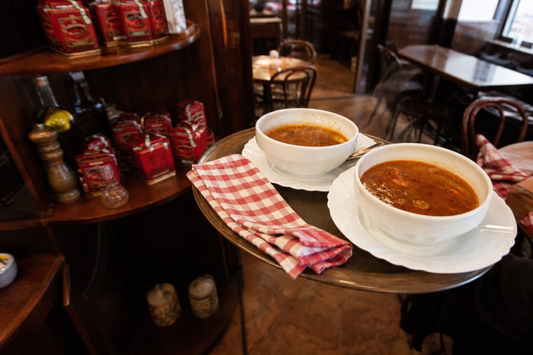 Small Budapest restaurant interior at lunchtime, red-checked napkins and worn bentwood chairs, a waiter's tray carrying two steaming bowls of goulash soup past shelves of paprika tins, warm tungsten light, no faces in focus